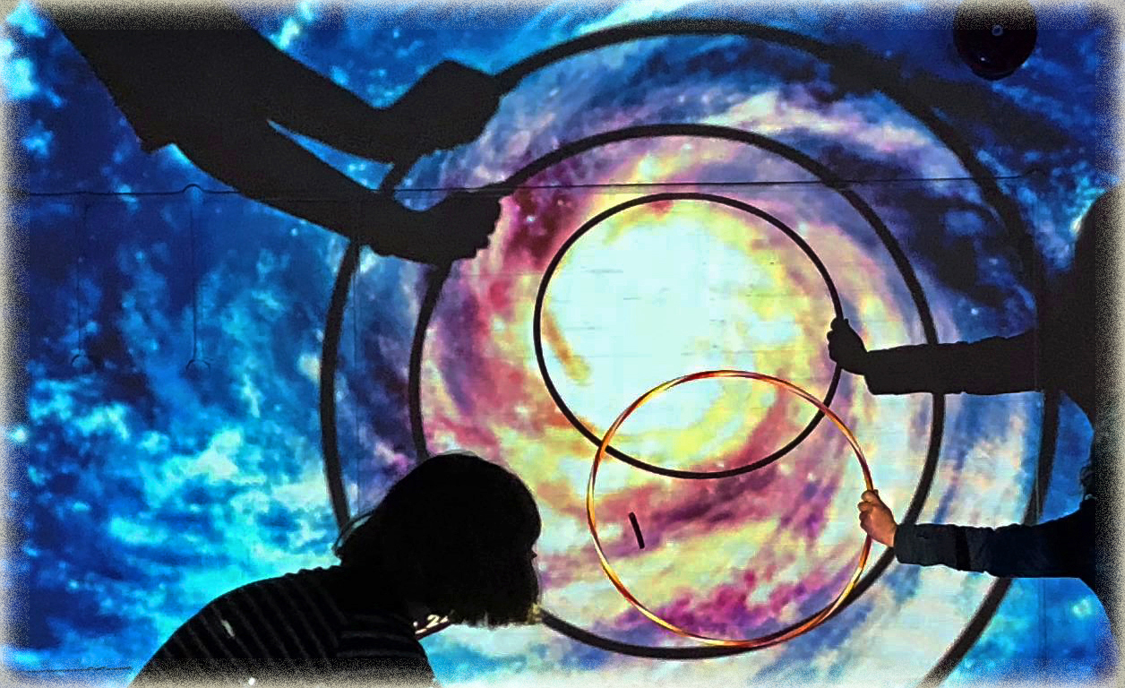 3 people in silhouette hold different sized hoops in front of a spacey galaxy projection