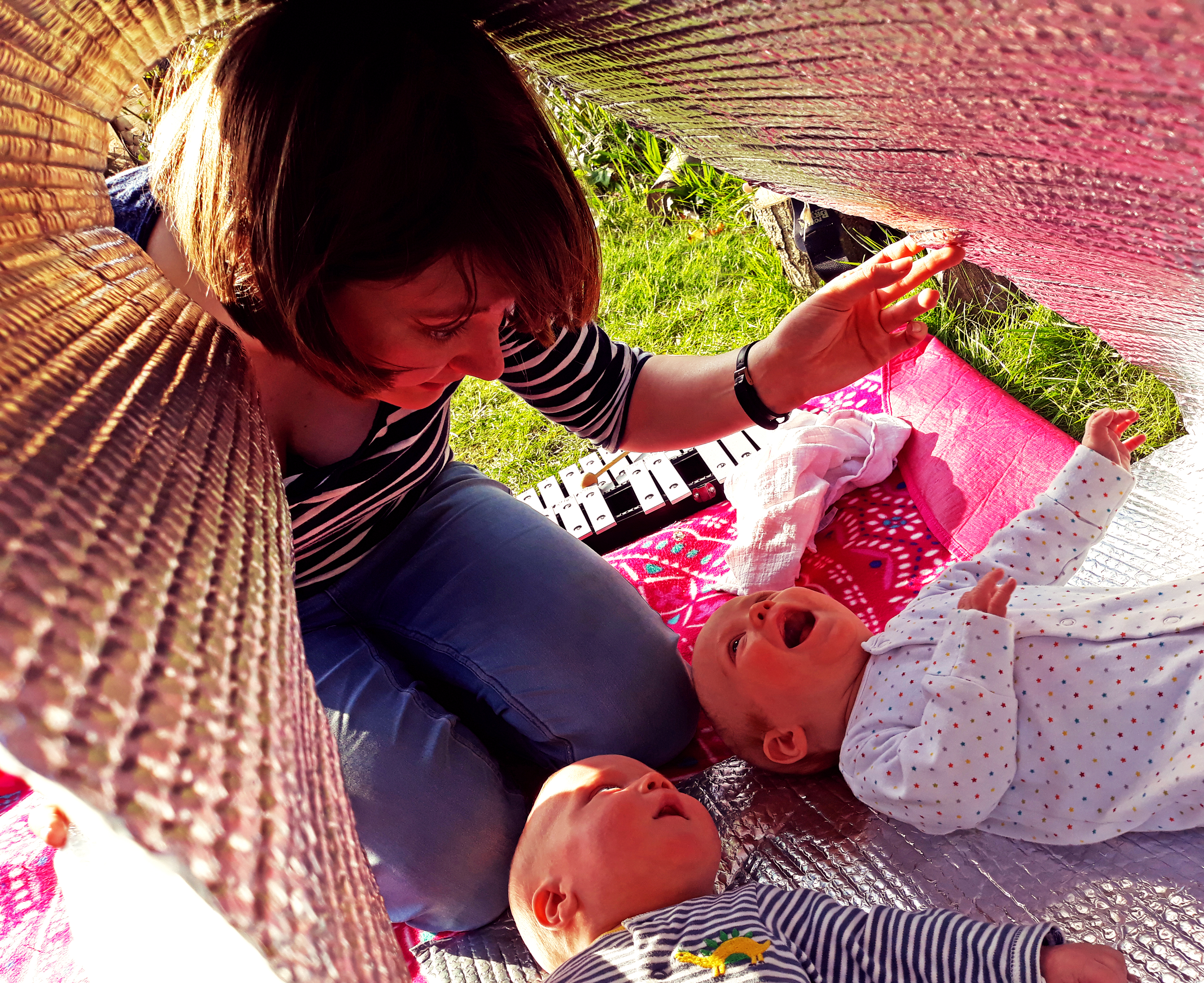 A young white woman with brown hair in a bob kneels in a sensory den, playing with the reflections of light on some bubbly silver material to the delight of one baby and cautious curiosity of another. In the background there is grass and a glockenspiel.