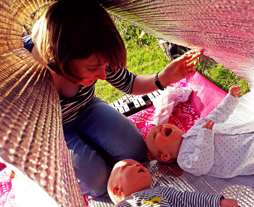 Two amazing and amazed babies lie on their backs in a cavern of silver reflective bubbly material, which is held up by a white woman with a bob. In the background there are grass and a glockenspiel.
