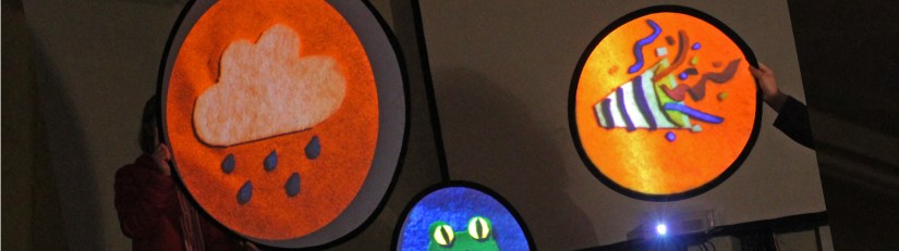 Three handheld circular projection screens showing large felt symbols: a weather-symbol rain cloud, a party-popper emoji and a frog emoji