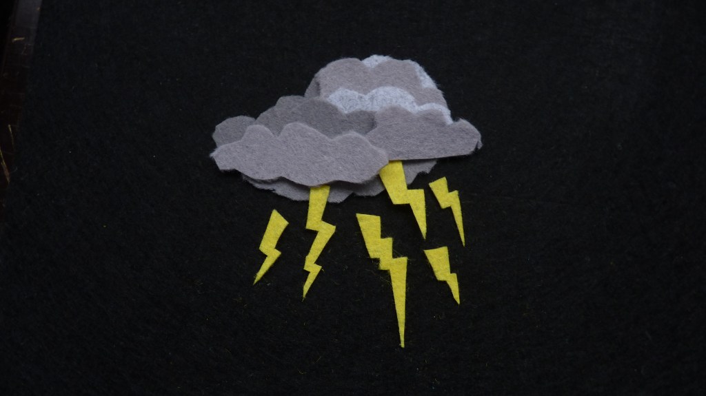 A multi-layered felt thundercloud in lots of shades of grey against a black felt background. The cloud has six jaggedy yellow zigzags of lightning coming out of it in different directions, some smaller, a couple of 'em whoppers. It looks quite scary and a bit angry.