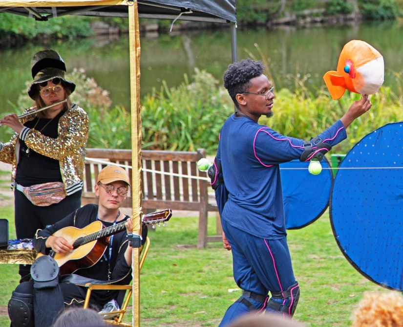 A photo of two white musicians and a young black puppeteer performing outdoors