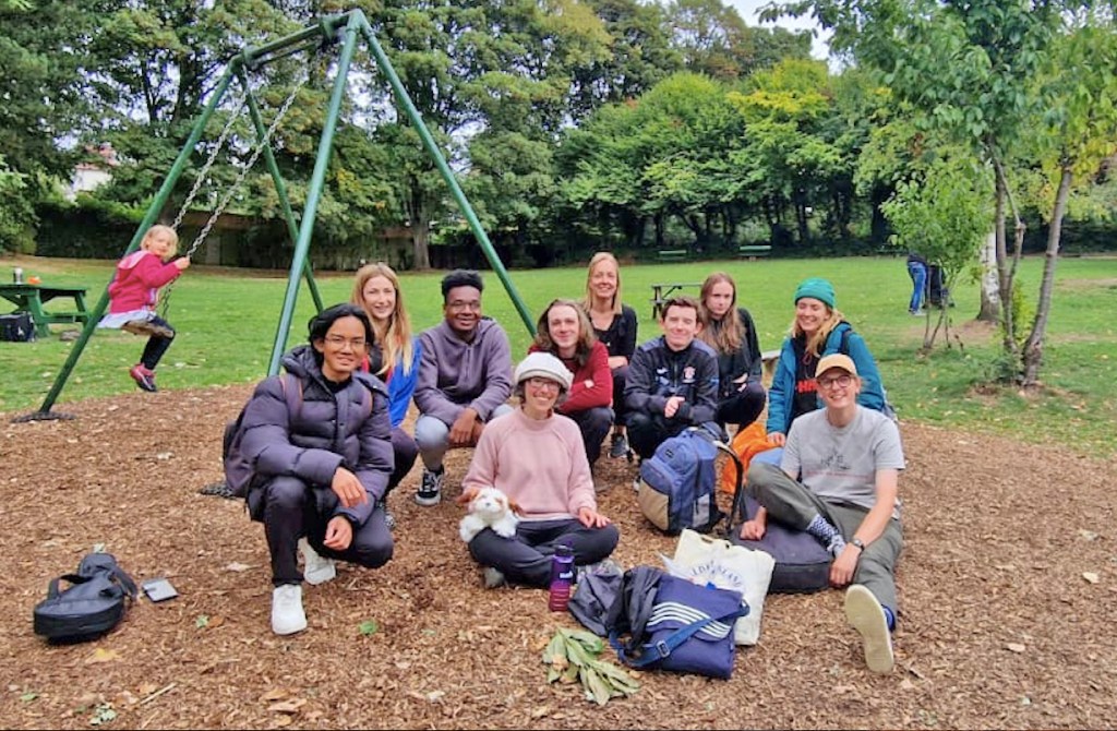 A team photo taken in a park: 10 people of varied ethnicity and gender from their teens to middle age sit on the ground while a little blonde-haired girl in a red coat watches from the swing behind them.