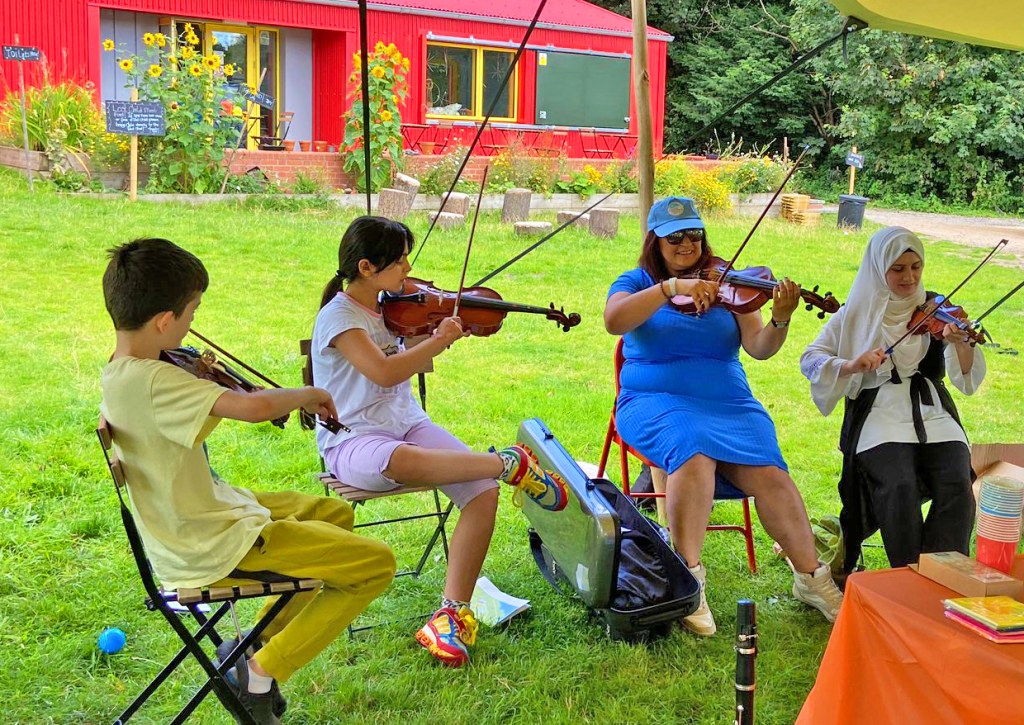 Our facilitator and 3 children play violin on the grass, with Red Shed and bundles of sunflowers in the background