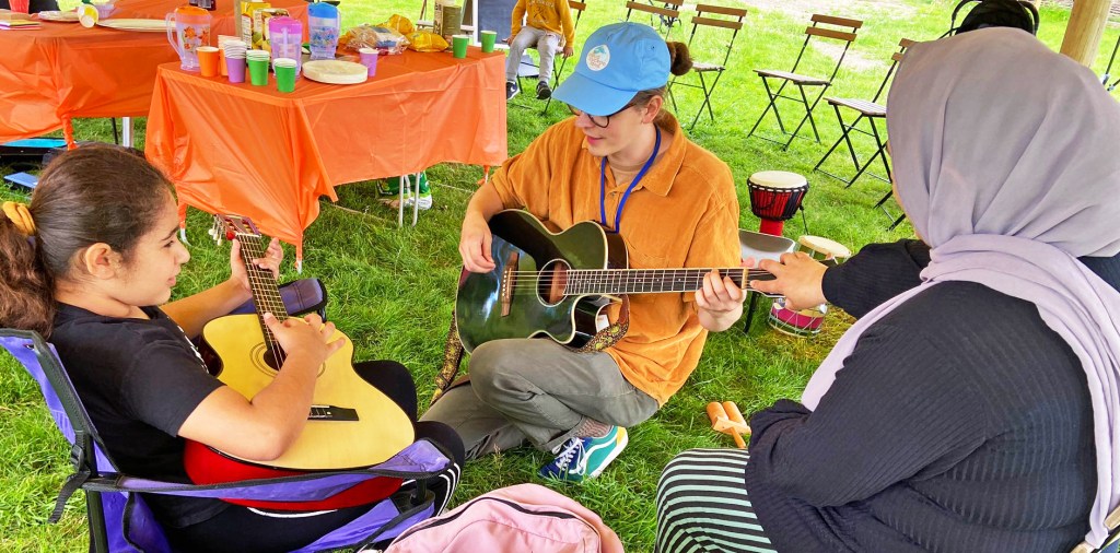 A young girl sits in a camping chair playing a small guitar, supported by a kneeling facilitator and her Mum