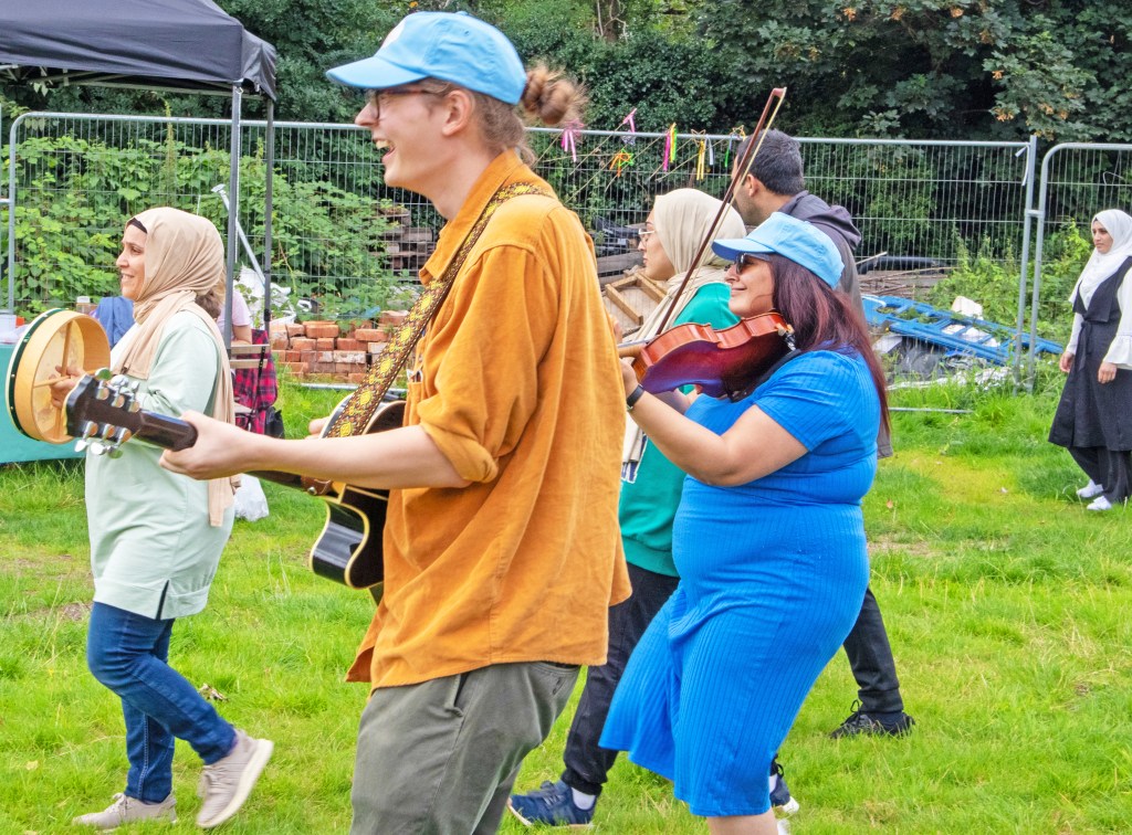 A group of children, parents and facilitators walk along playing music on drums, a guitar and a viola