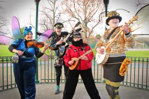Four people dressed in glittery, vintage-style insect costumes stand in a park playing violin (a dragonfly), guitar (a beetle), banjo (a bee) and mandolin (a ladybird)