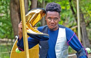 A photo of a young black man with glasses wearing a silver waistcoat: he is in a play, acting being on a phone made out of gold fabric.